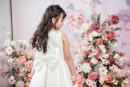 Young girl in a white dress standing in front of a floral arrangement with a light purple background