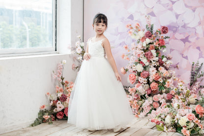 Young girl in a white dress standing in front of a floral arrangement and butterfly wall decoration.