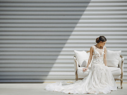 Woman in a white wedding dress sitting on a white chair against a gray wall.