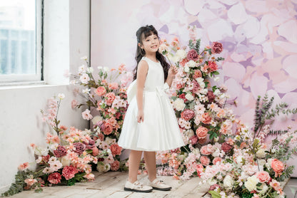 Young girl in a white dress standing among flowers with a decorative floral wall in the background