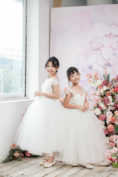 Two young girls in white dresses standing in front of a floral arrangement and a decorative backdrop.